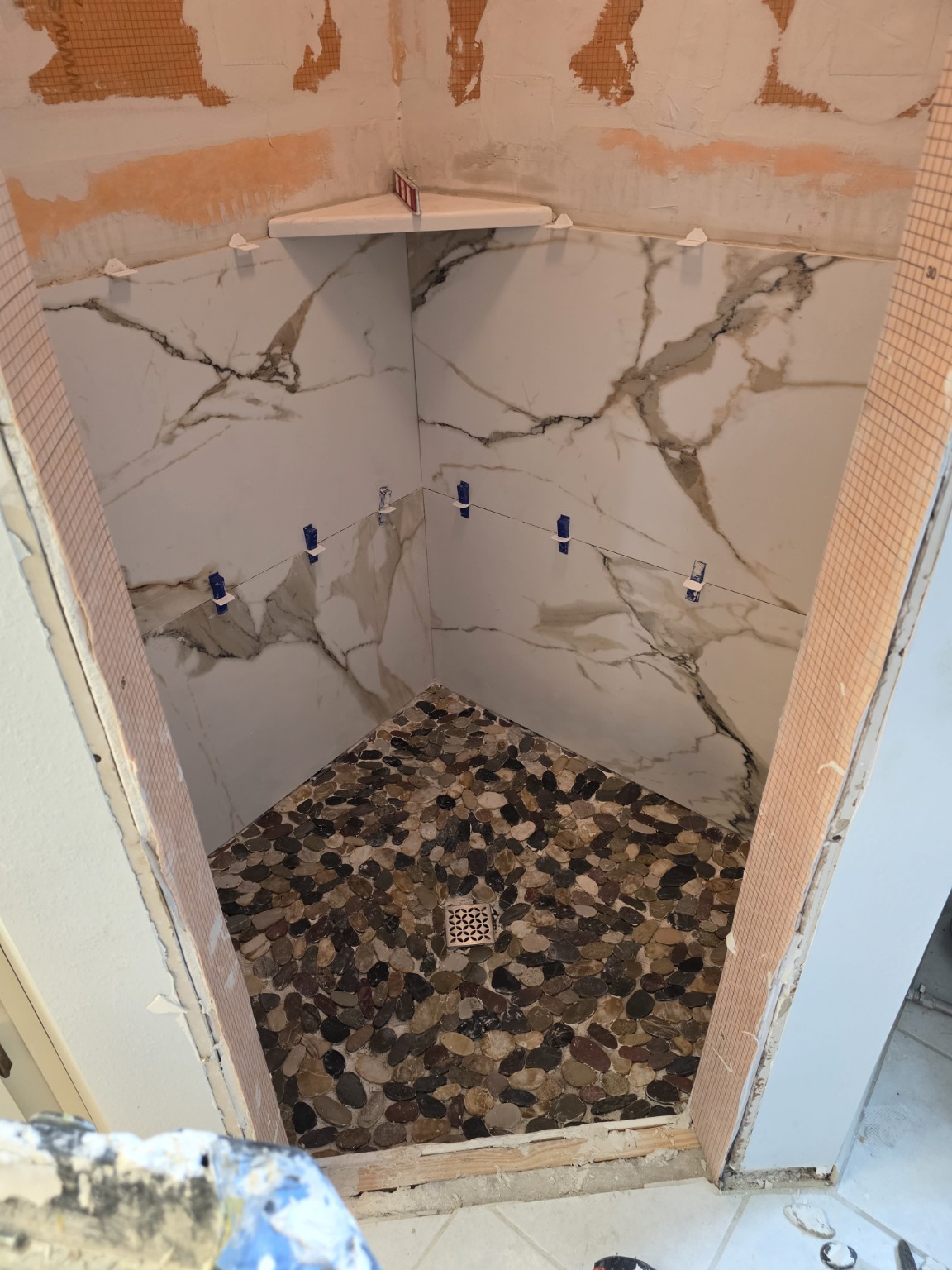 Marble-pattern lower shower wall tiles and corner shelf being installed above pebble floor